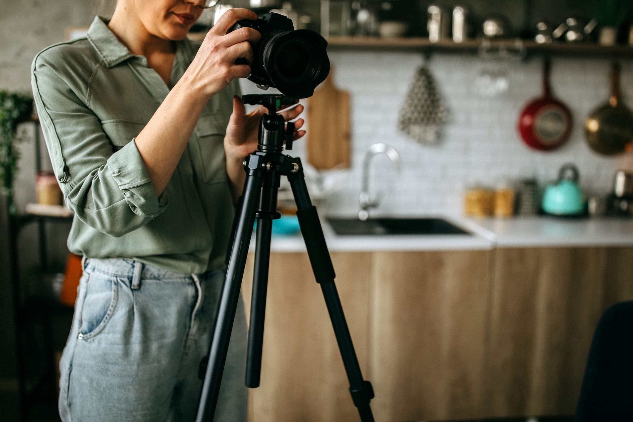 En kvinne justerer et kamera på stativ i et moderne kjøkken, forberedt til boligfotografering; bakgrunnen viser rene benkeplater, hengt kjøkkenutstyr og god belysning – ideelt for å fremheve interiørets detaljer.